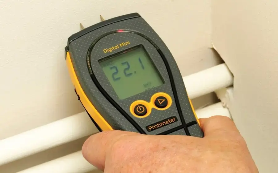 Hand holding a pin moisture meter against a wall above two pipes during a Glendora water damage inspection